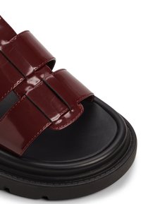 Burgundy patent leather sandal with thick black platform sole, wide straps featuring a woven design, and a smooth finish.