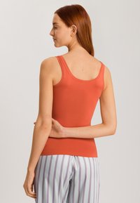 Top senza maniche, aderente, di colore terracotta, realizzato in tessuto elastico. Presenta una scollatura a barca e una texture liscia. Abbinato a pantaloni a righe.