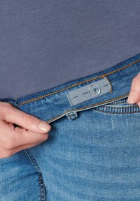 Světle modré džíny s výrazným oranžovým prošitím, s šedou látkovou visačkou se dvěma stehými linkami. Ruka držící pásek.