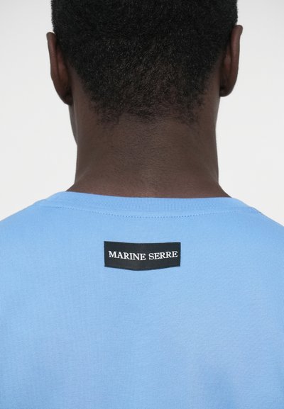 Svetlomodré bavlnené tričko s okrúhlym výstrihom a obdĺžnikovou čiernou nášivkou s bielym textom "MARINE SERRE" na zadnej strane.