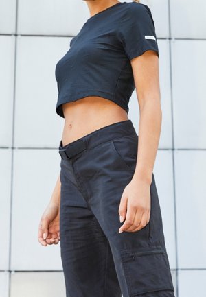 Personne portant un t-shirt noir court et un pantalon cargo noir taille haute avec une ceinture, debout devant un mur carrelé.