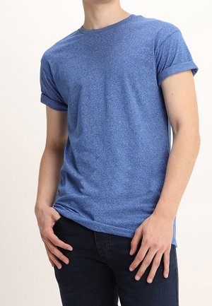 Niebieska koszulka z krótkim rękawem o teksturze melanżowej i podwiniętych mankietach, zestawiona z ciemnymi dżinsami. Krój jest swobodny i luźny.