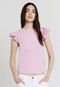 Top rosa a coste con maniche a farfalla bordate in pizzo, scollatura rotonda e vestibilità leggermente cropped. Il modello indossa pantaloni bianchi e ha i capelli lunghi.