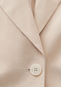Primo piano di un blazer beige su misura, con una texture sottile, rever affilati e un unico bottone bianco con quattro fori.