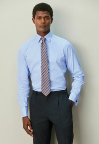 Hellblaues Hemd mit dezentem Karomuster, kombiniert mit dunklen Hosen. Hat lange Ärmel und eine pinkgestreifte Krawatte.