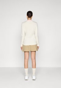 Pull en rib lisse crème avec manches longues, texture douce, col rond. Associé à une jupe plissée beige. Le modèle porte des baskets blanches et des chaussettes claires.