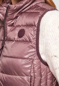 Chaleco acolchado de color púrpura con un cuello alto, que presenta un logo redondo y un ribete en contraste. Tejido suave y brillante con un patrón de costuras visible.