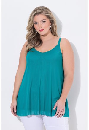 Femme aux longs cheveux blonds porte un haut sans manches bleu sarcelle et un pantalon blanc, debout devant un fond blanc, souriant doucement.