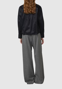 Kobieta o długich włosach, ubrana w czarną bluzkę z długim rękawem i szare szerokie spodnie w prążki, stoi tyłem na jednolitym tle.