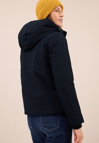 Giacca puffer blu navy con cappuccio, caratterizzata da un design trapuntato e polsini elasticizzati. Abbinata a un berretto in maglia giallo a coste e jeans blu.