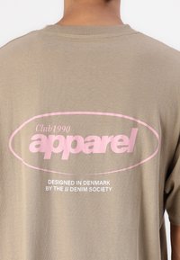 Camiseta de algodón verde oliva con un gráfico ovalado rosa que presenta el texto "Club1990 apparel" y un texto más pequeño en blanco debajo. Mangas cortas y cuello redondo.