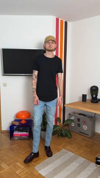 Fekete póló, világoskék farmernadrág és sötét cipő. Lazán illeszkedő öltözék, a karokon tetoválások láthatók, egy színes csíkos háttér előtt áll.