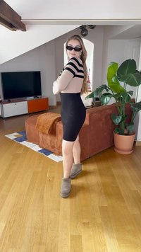 Melna ķermeni pieguļoša kleita ar neitralām svītrām augšpusē, gari mati, melni saulesbrilles, pelēki potīšu zābaki. Fona attēlā ir dīvāns un augi.