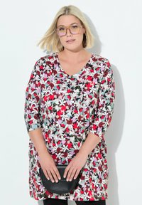 Bloemen tuniek met een V-halslijn, 3/4 mouwen, met een multicolore patroon van rood, roze en groen op een lichte achtergrond. Zwarte handtas.
