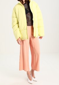 Chaqueta amarilla acolchada con cierre delantero, llevada sobre una camiseta de malla negra, combinada con pantalones de pierna ancha de color rosa claro y tacones blancos. Textura suave.