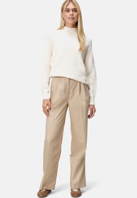 Cremefarbener gerippter Pullover mit hohem Kragen, kombiniert mit beigen, gefalteten Hosen. Der Look wird mit braunen Schnürschuhen abgerundet.