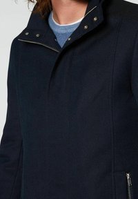 Chaqueta azul marino con superficie texturizada, cuello alto y botones metálicos. Dispone de un bolsillo con cremallera en el lateral para almacenamiento.