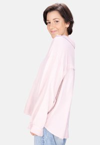 Light pink, oversized button-up shirt with a textured fabric, long sleeves, and a collar, displayed on a model with blue jeans.