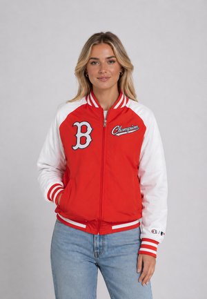 Femme aux cheveux blonds portant une veste zippée rouge et blanche avec les logos Boston "B" et Champion, debout avec une main dans la poche et un jean bleu clair.