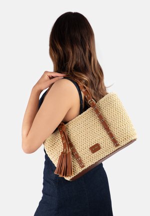 Žena s dugom smeđom kosom drži bež pletenu torbu za nošenje s smeđim kožnim remenima i resama preko ramena na bijeloj pozadini.