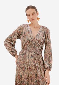 Šaty s dlouhým rukávem v tlumených barvách, s hlubokým výstřihem do V, nařaseným pasem a měkkou, texturovanou tkaninou s jemnými vzory.