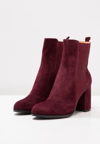 Stivaletti alla caviglia in suede bordeaux con un pannello in elastico a coste, punta affusolata e tacco spesso. Il design presenta una linguetta per indossarli facilmente.