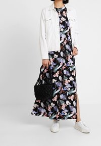 Dlouhé černé květinové šaty s růžovými a modrými vzory, doplněné bílou denimovou bundou. Doplňky zahrnují černou pletenou tašku a bílé tenisky.