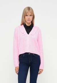 Ροζ πλεκτό cardigan με cropped σχέδιο, διαθέτοντας σχέδια καλωδίωσης, φαρδύ γιακά και μπροστινό κλείσιμο με κουμπιά. Φορεμένο επάνω από σκούρα τζιν.