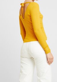 Vrouw in een mosterdgele ribbed top met lange mouwen en een strikdetail op de rug, gecombineerd met witte high-waisted broeken, tegen een effen achtergrond.