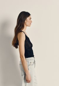 Young woman with long brown hair wearing a black sleeveless top and light blue jeans, standing in profile against a plain background.