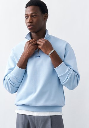 Νέος άνδρας με ανοιχτό γαλάζιο φούτερ με φερμουάρ να διορθώνει τον γιακά, φοράει ασημένιο βραχιόλι, στέκεται μπροστά σε απλό λευκό φόντο.