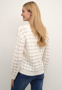 Maglione bianco lavorato a maglia con un motivo a punto aperto, scollatura tonda e polsini a costine. Abbinato a jeans in denim blu.