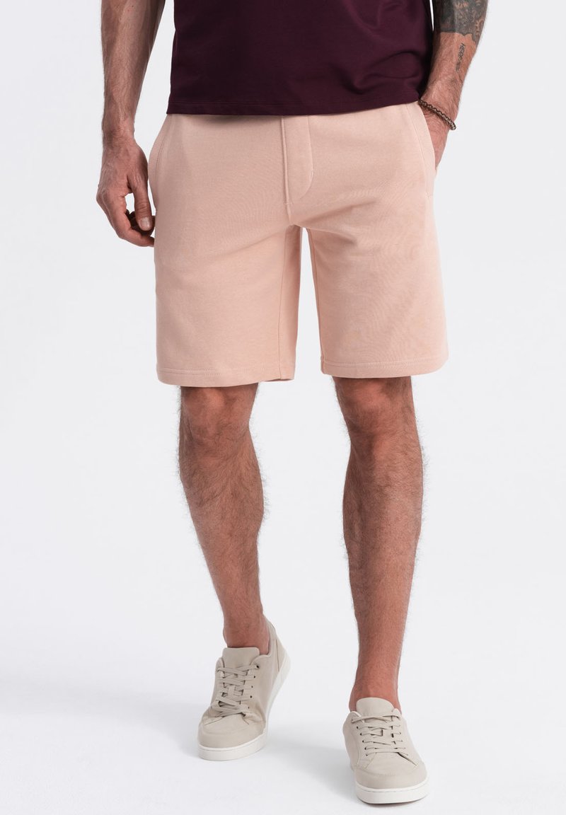 Světlé růžové šortky z měkké tkaniny s elastickým páskem a bočními kapsami, doplněné o světlé šedé sneakers a tmavý top.
