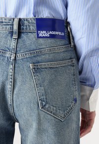 Bakåtsyn av en person som bär en blårandig skjorta och ljusblå Karl Lagerfeld jeans med logotyppatch ovanför den högra bakfickan.