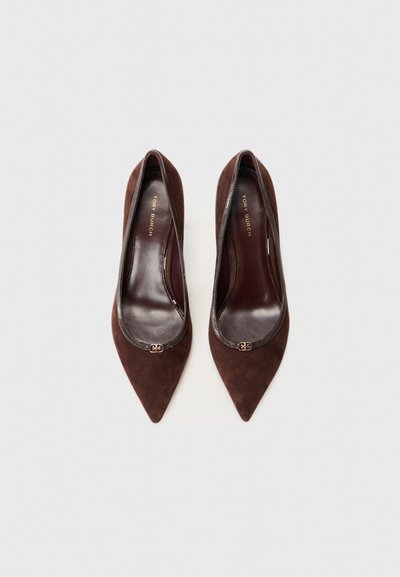 Brown suede pointed-toe pumps with a dark leather trim, featuring a gold logo accent on the front and a smooth interior lining.