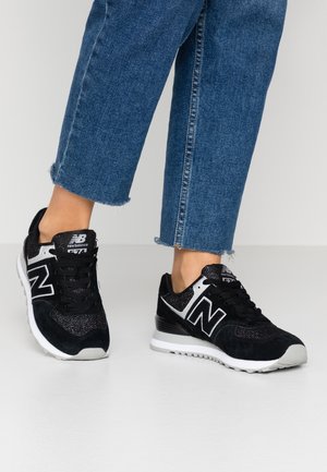 Osoba nosí černobílé tenisky New Balance 574 spojené s modrými džínami s třepenými lemy na bílém jednobarevném pozadí.