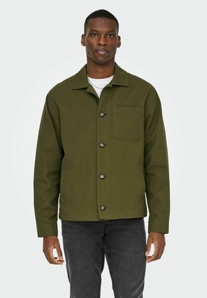 Mand iført en olivengrøn knap-op jakke med brystlomme, hvid undertrøje og mørkegrå jeans, stående mod en ensfarvet baggrund.