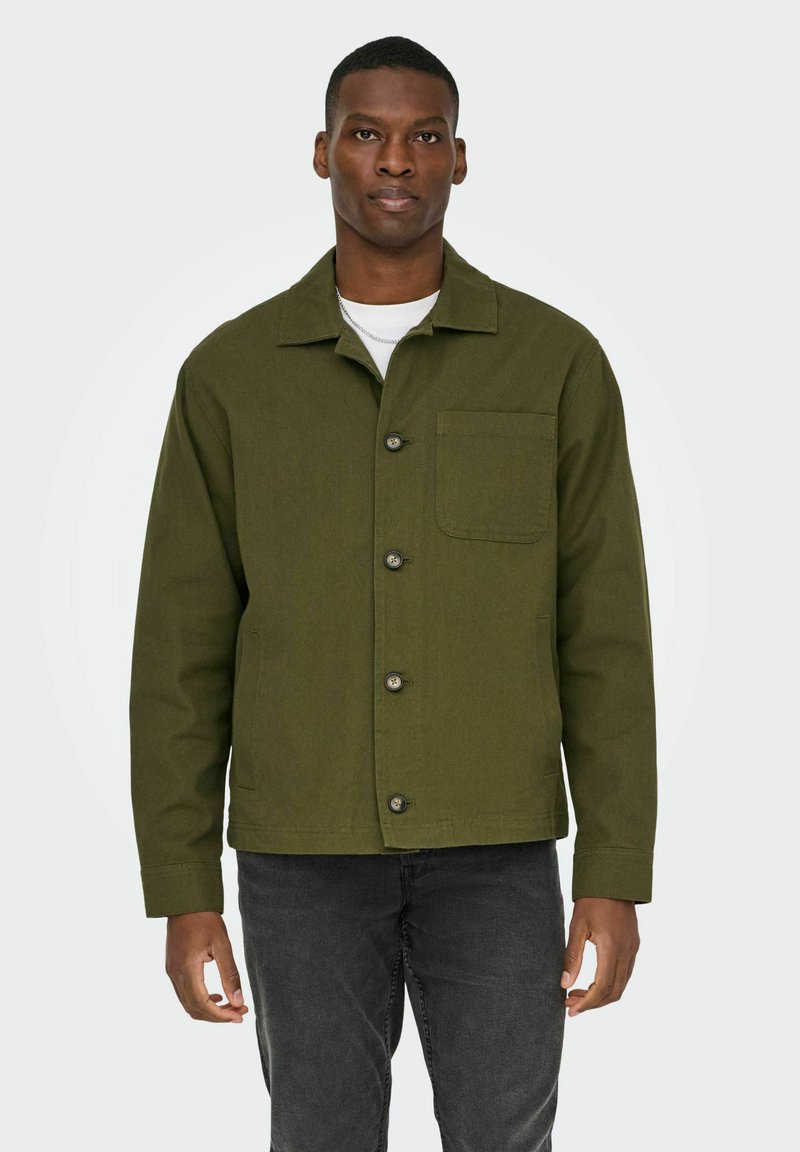 Moški, oblečen v olivno-zeleno jakno z gumbi in žepom na prsih, belo spodnjo majico in temno sive kavbojke, stoji pred enobarvnim ozadjem.