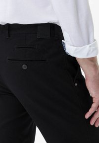 Pantaloni neri con una texture liscia, tasche posteriori con bottone e dettagli di cucitura sottili. L'etichetta attaccata sulla cintura è visibile.