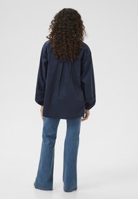 Temno moder oversize plašč z dolgimi, voluminoznimi rokavi in sproščenim krojem, v kombinaciji z razširjenimi denim hlačami. Gladka tekstura in sproščen dizajn.