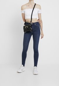 Bolso bandolera negro con una hebilla dorada, llevado sobre un top corto blanco de hombros descubiertos y jeans ajustados azul oscuro. Zapatillas blancas completan el conjunto.