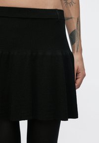 Čierna pletená sukňa s textúrovanou látkou, vybavená priliehavým pásom a rozšíreným lemom, zobrazená s rukou na boku pre mierku.