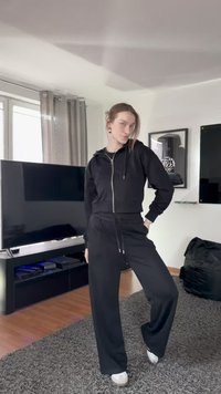 Czarna bluza z kapturem na zamek, o krótkim kroju, z przednimi kieszeniami, zestawiona z luźnymi czarnymi spodniami dresowymi. Sceneria to nowoczesne wnętrze z szarymi dywanami.