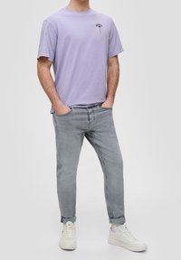 Camiseta de algodón de color púrpura claro con mangas cortas y un pequeño diseño floral en el pecho, combinada con jeans gris claro y zapatillas blancas.