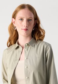 Light green button-up shirt with a classic collar, and subtle texture. Worn over a beige tank top, features a small logo on the chest.