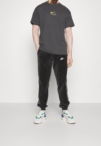 Šedé bavlněné tričko Nike se žlutým logem, v kombinaci s černými sametovými tepláky s bílým logem a bočními pruhy, doplněné barevnými teniskami.