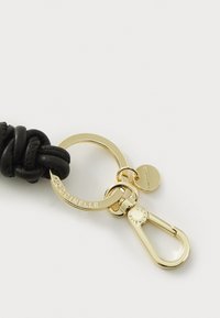 Portachiavi in pelle nera con design annodato, dotato di un anello e gancio in metallo dorato, logo inciso e un piccolo tag circolare appeso.