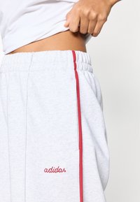 Ανοιχτό γκρι φόρμα με κόκκινες λωρίδες στο πλάι και κεντημένο λογότυπο «adidas». Το ύφασμα έχει απαλή υφή και ελαστική μέση.
