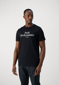 Camiseta de algodón negra de manga corta con el logo "Peak Performance" y "Est. 1986" en texto blanco en la parte delantera.
