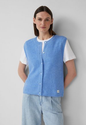 Gilet lavorato a maglia blu chiaro con un motivo testurizzato, scollatura tonda e bottoni frontali, indossato sopra una camicia bianca. Presenta un piccolo logo all'orlo.
